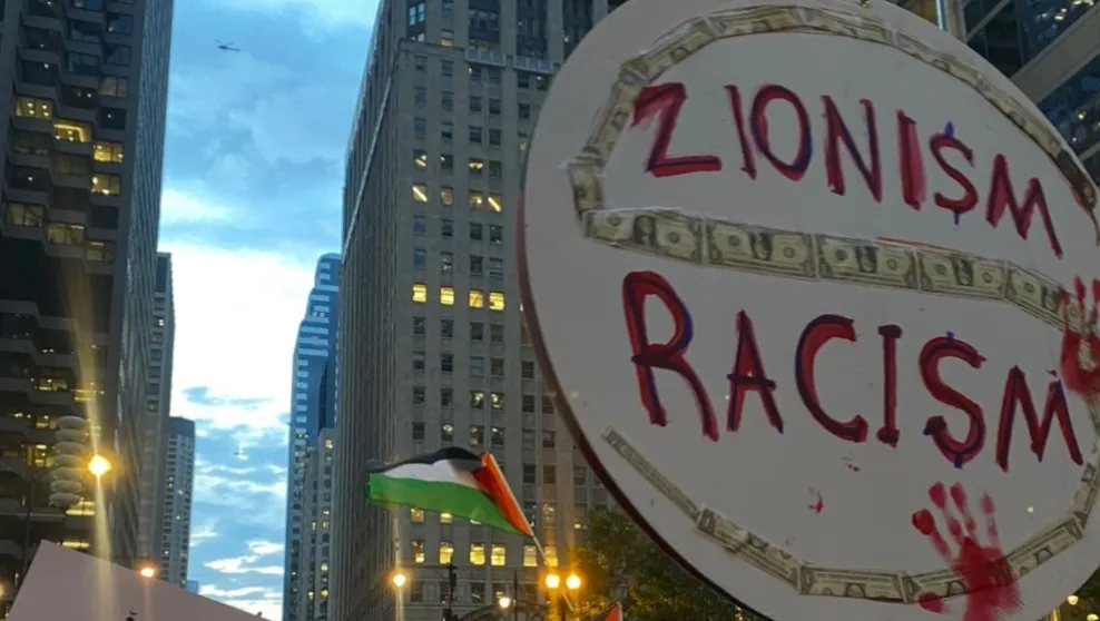 Photo: Chicago, Ill. – At a rally in October, a protester held a sign comparing Zionism and racism. The sign also contains a drawing of a snake formed from dollar bills.